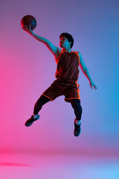 Joven hombre enérgico jugando baloncesto aislado en gradiente rosa azul estudio fondo en luz de neón. Juventud, hobby, movimiento, actividad, conceptos deportivos. — Foto de Stock