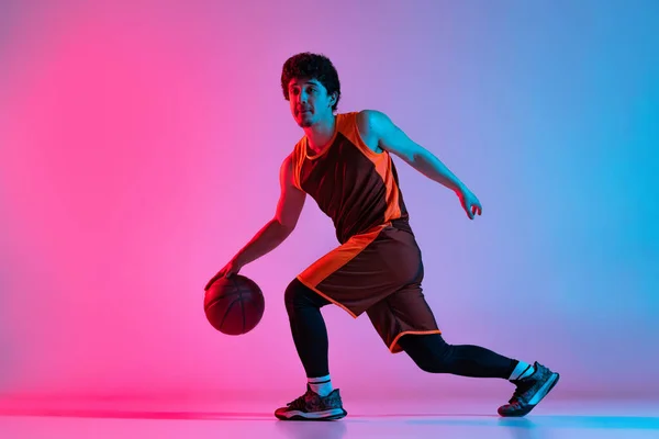 Uomo sportivo che gioca a basket isolato su sfondo rosa gradiente studio blu in luce al neon. Giovani, hobby, movimento, attività, concetti sportivi. — Foto Stock