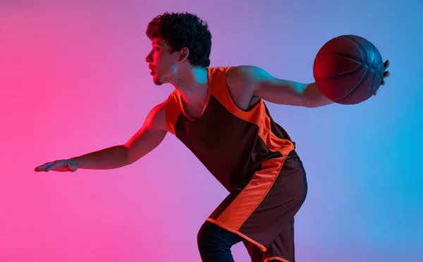 Giovane uomo energico che gioca a basket isolato su sfondo rosa gradiente studio blu in luce al neon. Giovani, hobby, movimento, attività, concetti sportivi. — Foto Stock