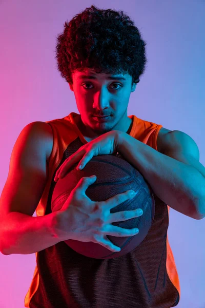 Portrait of young muscle man with basketball ball looking at camera isolated on gradient pink blue studio background in neon light. Youth, hobby, sport concepts.