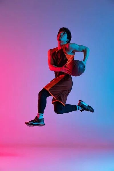 Homem esportivo jogando basquete isolado no fundo do estúdio gradiente rosa azul em luz de néon. Juventude, hobby, movimento, atividade, conceitos de esporte. — Fotografia de Stock