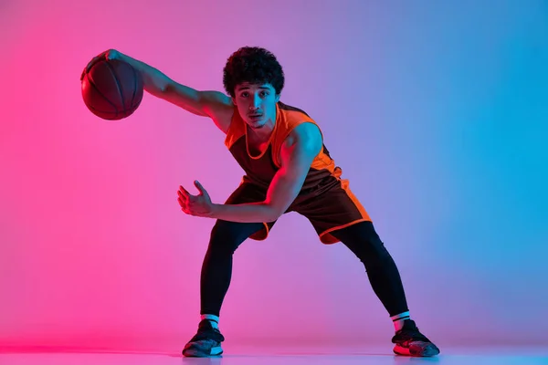 Jovem homem enérgico jogando basquete isolado no fundo do estúdio gradiente rosa azul em luz de néon. Juventude, hobby, movimento, atividade, conceitos de esporte. — Fotografia de Stock