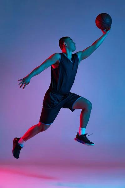 Joven hombre deportivo jugando baloncesto aislado en fondo estudio azul en luz de neón. Juventud, hobby, movimiento, actividad, conceptos deportivos. —  Fotos de Stock