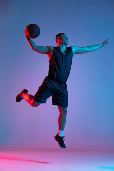 Joven deportista, jugador de baloncesto saltando con pelota aislada sobre fondo de estudio azul en luz de neón. Juventud, hobby, movimiento, actividad, conceptos deportivos. —  Fotos de Stock