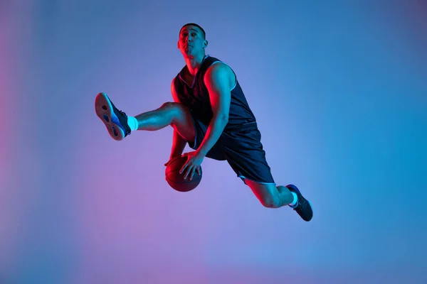 Młody sportowiec grający w koszykówkę na niebieskim tle studia w neonowym świetle. Młodzież, hobby, ruch, aktywność, koncepcje sportowe. — Zdjęcie stockowe