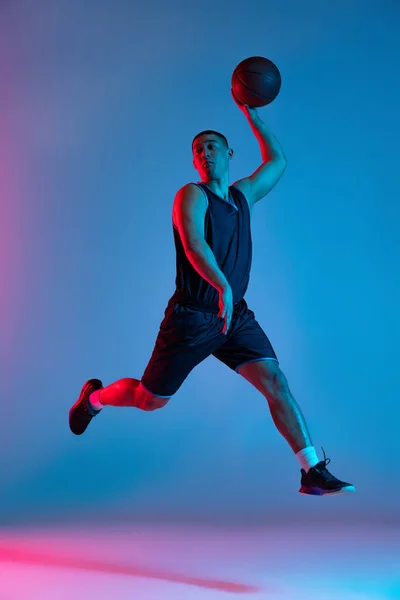 Jonge sportieve man, basketbalspeler springen met bal geïsoleerd op blauwe studio achtergrond in neon licht. Jeugd, hobby, beweging, activiteit, sportconcepten. — Stockfoto