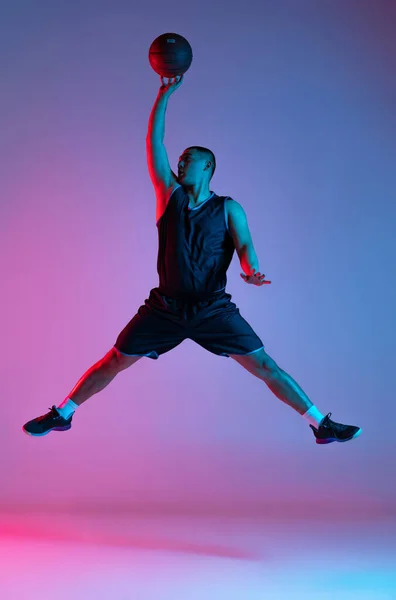 Młody sportowiec, koszykarz skaczący z piłką odizolowaną na niebieskim tle studia w neonowym świetle. Młodzież, hobby, ruch, aktywność, koncepcje sportowe. — Zdjęcie stockowe