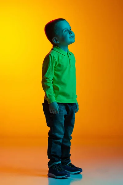 Full-length portret van charmante kleine jongen poseren geïsoleerd op gele studio backgroud in neon licht. Concept van kinderemoties, gezichtsuitdrukking, actie, kindertijd — Stockfoto