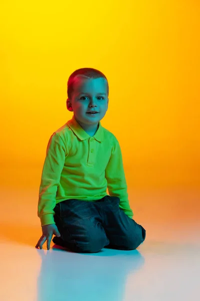 Studio shot of cute little boy, kid sitting on floor isolated on yellow studio backgroud in neon light. Concept of child emotions, facial expression, childhood — Stock Photo, Image