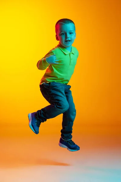 Full-length portret van charmante kleine jongen poseren geïsoleerd op gele studio backgroud in neon licht. Concept van kinderemoties, gezichtsuitdrukking, actie, kindertijd — Stockfoto