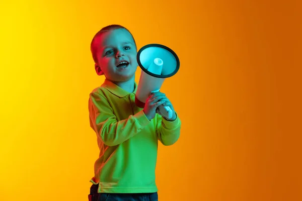 Un petit garçon dans des vêtements de style décontracté tenant mégaphone isolé sur fond de studio jaune dans la lumière au néon. Concept d'émotions de l'enfant, expression faciale, enfance — Photo