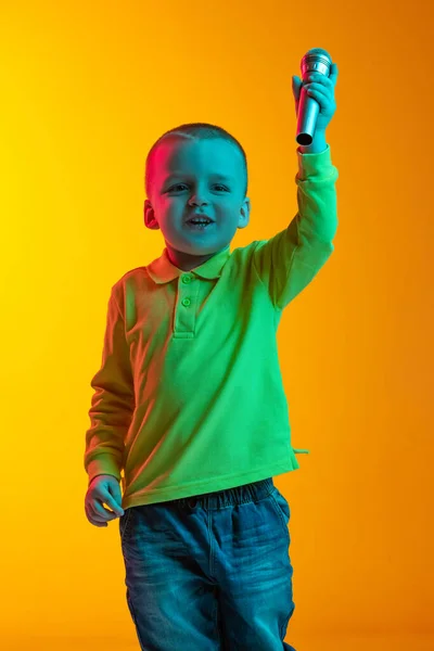 Porträtt av liten pojke i avslappnad stil kläder sjunger vid mikrofon isolerad på gul studio backgroud i neon ljus. Begreppet barns känslor, ansiktsuttryck, barndom — Stockfoto