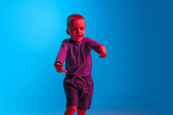 Retrato de niño pequeño, niño, niño en edad preescolar aislado en el fondo del estudio azul en luz de neón rojo. Concepto de emociones infantiles, expresión facial, infancia — Foto de Stock