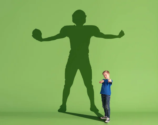 Εννοιολογική εικόνα με το μικρό παιδί να ονειρεύεται τη μελλοντική αθλητική καριέρα και τη σκιά του αρσενικού αμερικανού ποδοσφαιριστή σε πράσινο φόντο. — Φωτογραφία Αρχείου