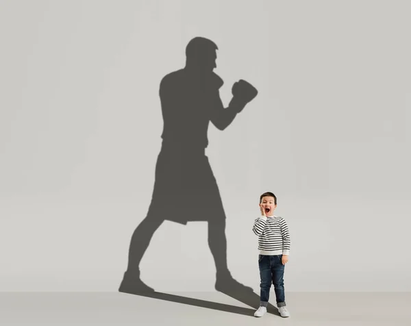Obraz koncepcyjny z chłopcem i cieniem sportowego boksera, mistrz na szarym tle. Dzieciństwo, marzenia, wyobraźnia, koncepcja edukacji. — Zdjęcie stockowe