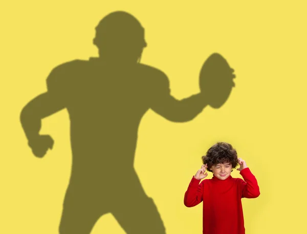 Konceptualny obraz z małym dzieckiem marzącym o przyszłej karierze sportowej i cieniu amerykańskiego piłkarza na żółtym tle. — Zdjęcie stockowe