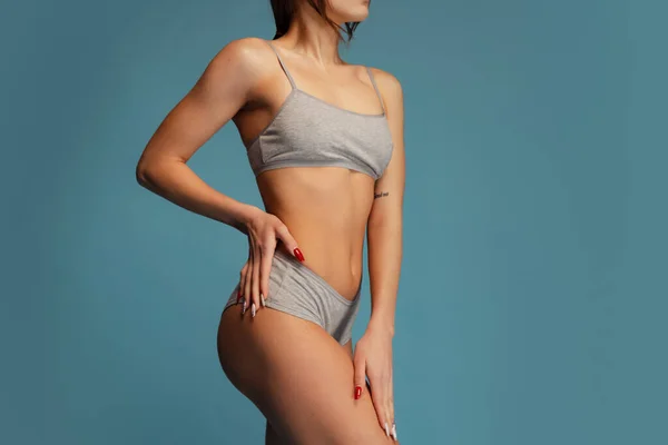 Обрізаний студійний знімок тонкого спортивного жіночого тіла в сірій бавовняній білизні, що позує ізольовано на синьому фоні. Концепція фітнесу — стокове фото