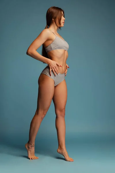 Полноразмерный портрет красивой женщины с идеальным спортивным телом, позирующим в сером хлопчатобумажном нижнем белье, изолированном на синем фоне студии — стоковое фото