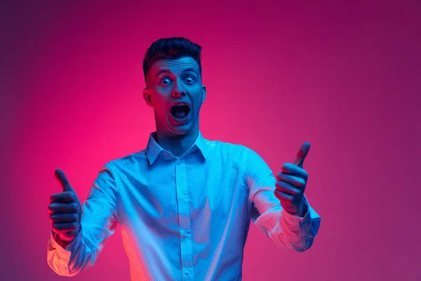 Aufgeregt junger stilvoller Mann, Student isoliert über magentafarbenem Studiohintergrund in blauem Neonlicht. Konzept der Emotionen, menatl Gesundheit, Nachrichten — Stockfoto