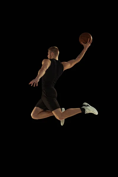 Dynamisch portret van jongeman, basketbalspeler in zwart uniform training geïsoleerd op donkere achtergrond. Resultaten, sportcarrière, bewegingsconcepten. — Stockfoto