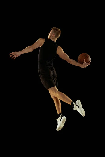 Dynamisches Porträt eines jungen Mannes, Basketballspieler in schwarzer Uniform, der isoliert auf dunklem Hintergrund springt. Erfolge, Sportkarriere, Bewegungskonzepte. — Stockfoto