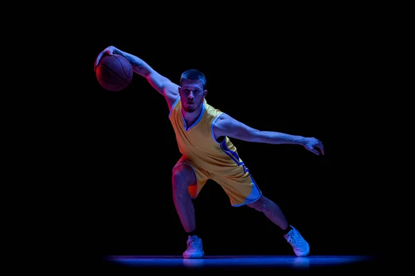 Portret van sportieve man, professionele basketbalspeler die basketbal speelt geïsoleerd op donkere achtergrond in neon licht. Resultaten, sportcarrière, bewegingsconcepten. — Stockfoto
