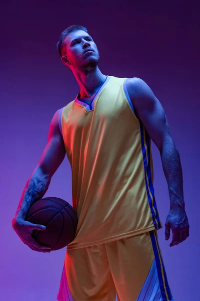 Studyjne ujęcie umięśnionego mężczyzny, koszykarza stojącego z piłką odizolowaną na fioletowym tle w neonowym świetle. Cele, sport, ruch, koncepcje aktywności. — Zdjęcie stockowe