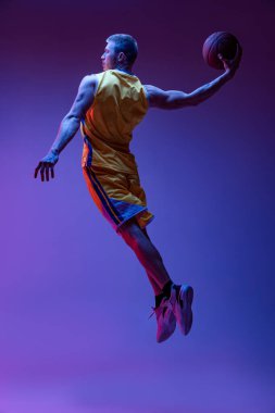 Sportif bir adamın dinamik portresi, neon ışıkta mor arka planda izole edilmiş top ile basketbol oyuncusu eğitimi. Hedefler, spor, hareket, aktivite kavramları.