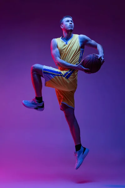 Studio shot of muscled man, basketball player training with ball isolated on purple background in neon light. Goals, sport, motion, activity concepts. — Stock Photo, Image