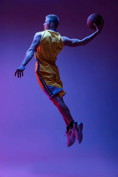 Portrait dynamique d'homme sportif, entraînement de joueur de basket-ball avec balle isolée sur fond violet sous la lumière du néon. Objectifs, sport, mouvement, concepts d'activité. — Photo