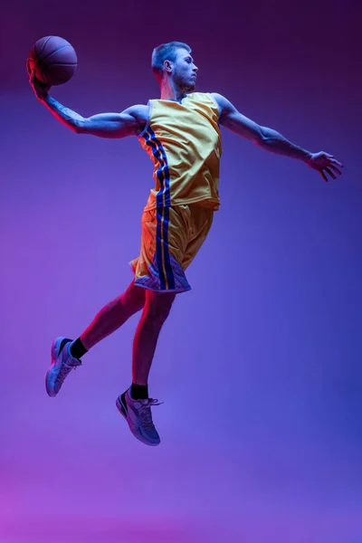 Retrato dinâmico de homem esportivo, treinamento de jogador de basquete com bola isolada em fundo roxo em luz de néon. Objetivos, esporte, movimento, conceitos de atividade. — Fotografia de Stock