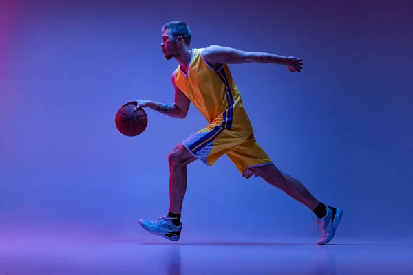 Studioaufnahme eines muskulösen Mannes, Basketballspieler beim Training mit Ball auf violettem Hintergrund in Neonlicht. Ziele, Sport, Bewegung, Aktivitätskonzepte. — Stockfoto