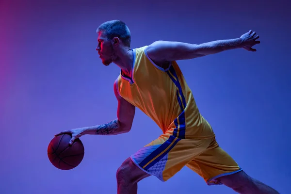 Studioaufnahme eines muskulösen Mannes, Basketballspieler beim Training mit Ball auf violettem Hintergrund in Neonlicht. Ziele, Sport, Bewegung, Aktivitätskonzepte. — Stockfoto