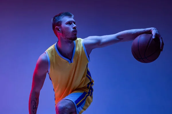 Young man, professional basketball player training with ball isolated on purple background in neon light. Goals, sport, motion, activity concepts.