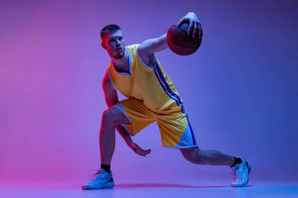 Junger Mann, professioneller Basketballspieler, der mit Ball auf lila Hintergrund im Neonlicht trainiert. Ziele, Sport, Bewegung, Aktivitätskonzepte. — Stockfoto