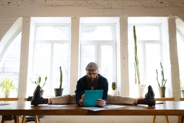 Joven barbudo, empleado de oficina divirtiéndose, haciendo yoga sobre mesa de madera en oficina moderna a la hora del trabajo con gadgets. Concepto de negocio, estilo de vida saludable, deporte, hobby — Foto de Stock