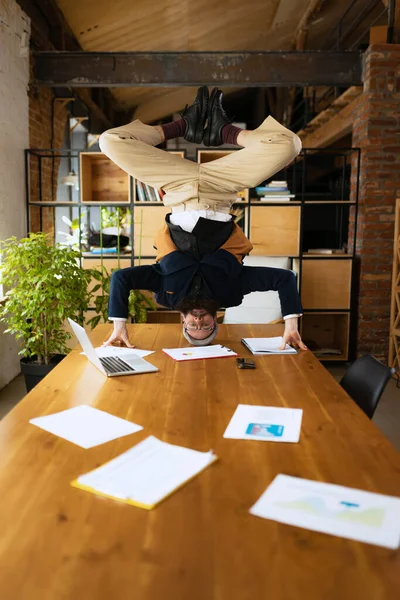 Joven barbudo, empleado de oficina divirtiéndose, haciendo yoga sobre mesa de madera en oficina moderna a la hora del trabajo con gadgets. Concepto de negocio, estilo de vida saludable, deporte, hobby — Foto de Stock