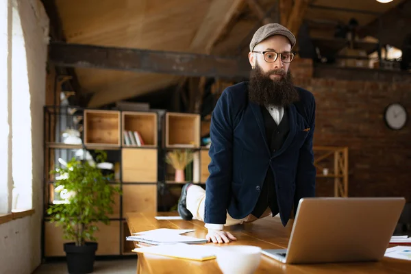 Un hombre, empleado de oficina que usa ropa de estilo empresarial que se divierte, haciendo yoga en la mesa de madera en la oficina moderna a la hora del trabajo con gadgets. Concepto de deporte, hobby — Foto de Stock