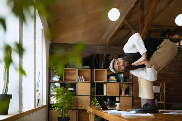 Tiempo de descanso. Joven barbudo, oficinista haciendo yoga sobre mesa de madera en oficina moderna a la hora del trabajo con gadgets. Concepto de negocio, estilo de vida saludable, deporte, hobby — Foto de Stock