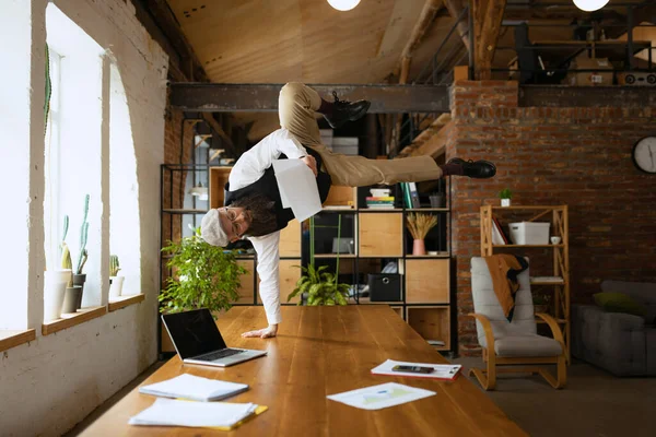 Joven barbudo, empleado de oficina divirtiéndose, haciendo yoga sobre mesa de madera en oficina moderna a la hora del trabajo con gadgets. Concepto de negocio, estilo de vida saludable, deporte, hobby — Foto de Stock
