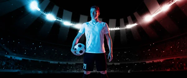 Un futbolista profesional, jugador de fútbol en kit de fútbol de pie con pelota sobre el estadio nocturno oscuro con linternas de fondo. — Foto de Stock