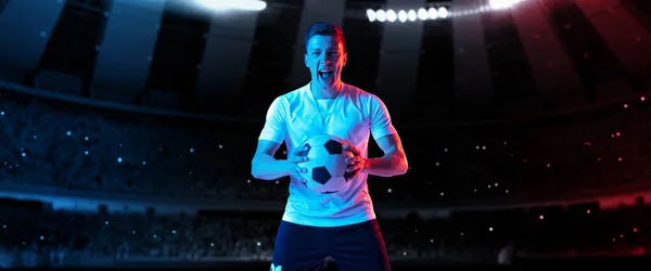 Win, goal. One professional soccer, football player in football kit standing with ball over dark night stadium with flashlights background. — Stock Photo, Image