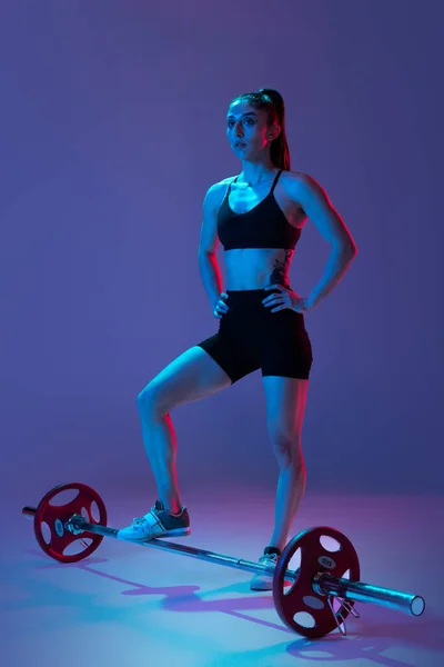 Πορτρέτο της μυώδης γυναίκα στην εκπαίδευση αθλητικών ενδυμάτων με ένα barbell απομονώνονται σε μωβ φόντο σε νέον φως. Αθλητισμός, άρση βαρών έννοια — Φωτογραφία Αρχείου