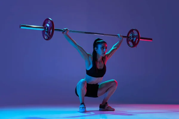 ネオンの光の中で紫の背景に隔離された鐘とスポーツウェアの訓練で筋肉質の女性の肖像画。スポーツ、重量挙げの概念 — ストック写真