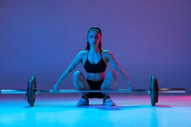 Spor giyim eğitiminde kaslı bir kadının portresi. Mor arka planda, neon ışıkta izole edilmiş bir halterle. Spor, halter konsepti