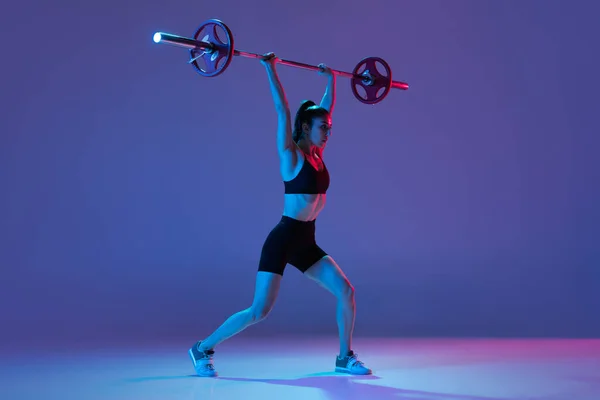 Retrato de mujer musculosa en entrenamiento de ropa deportiva con una barra aislada sobre fondo púrpura en luz de neón. Deporte, concepto de levantamiento de pesas —  Fotos de Stock