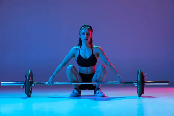 Портрет мускулистой женщины в спортивной форме, тренирующейся с штангой, изолированной на фиолетовом фоне в неоновом свете. Спорт, концепция тяжелой атлетики — стоковое фото