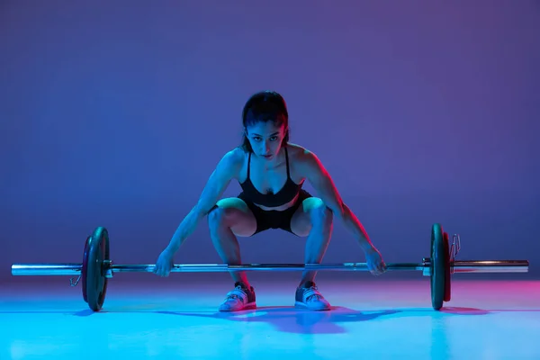 Retrato de mulher musculada em treinamento de sportswear com uma barra isolada em fundo roxo em luz de néon. Esporte, conceito de levantamento de peso — Fotografia de Stock