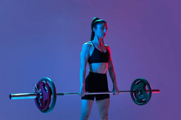 Retrato de mulher musculada em treinamento de sportswear com uma barra isolada em fundo roxo em luz de néon. Esporte, conceito de levantamento de peso — Fotografia de Stock