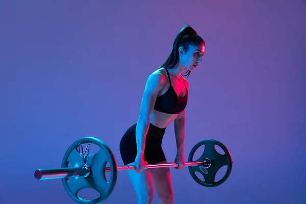 Estúdio tiro de atleta feminina, mulher esportiva posando com barra isolada em fundo roxo em luz de néon. Esporte, beleza, conceito de força — Fotografia de Stock